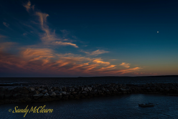 Crescent moon in top right and wispy orange clouds to the left over a breakwater in partial silhouette.