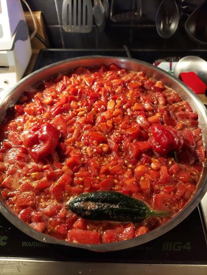 Chopped tomatoes, sugar, salt, and spices simmering in a pot. Floating in the tomatoes are 2 habanero and 1 jalapeño pepper. The way they are arranged makes it look like a face with the habaneros for eyes and the jalapeño a horizontal mouth