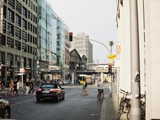 People are crossing a busy street, cars have stopped for the red light. A yellow tram car is stopped a little further back, and even further, an ICE train is coming into the scene on the train tracks above the street. The scene takes place in Berlin Friedrichstraße.