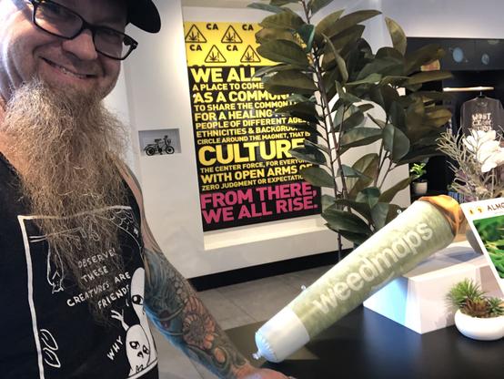 Standing next to an inflatable joint that reads "weedmaps" on the side. There is a very bright yellow and red lettered poster on the wall behind me.