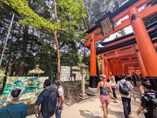 There are tourists in the middle.  There is a red Torii gate in the upper right corner.  This is on a path in the woods.