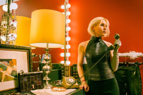 Photo de studio mise en scène, un décors intérieur de salle cosy retro-années folles contraste avec la tenue rétro-futriste du personnage personnage féminin apparent. Elle tient l'écouteur d'un ancien téléphone en bakélite - mais le tient loin de son oreilles. Elle n'écoute pas et a l'air totalement absente