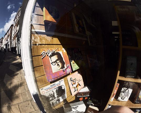 This image shows a shop window reflecting a busy street in York. The main focus is a colorful vinyl record cover featuring a man with glasses. The sunlight creates warm tones that highlight the textures of the shop's display and the surrounding buildings.