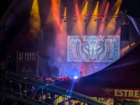El último grupo del festival: Pain of Salvation, se ve en la imagen de la pantalla del escenario el nombre y una imagen de la cabeza de frente de un animal con un símbolo en la frente y muchas líneas radiando hacia fuera.
El escenario con luces rojas y amarillas, una esquina de la carpa del bar, muchísima gente delante del escenario y una de las estructuras metálicas delante de la cámara.