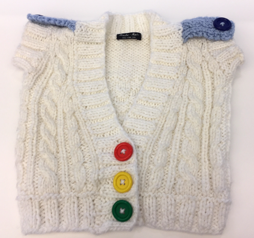 A custom-designed, white, cabled-knit, sweater-vest for a child is pictured flat on a white background. It has a tag sewn in at the neck, and two blue epaulets with blue buttons on the shoulders. The large three buttons are top to bottom: red, yellow, green.