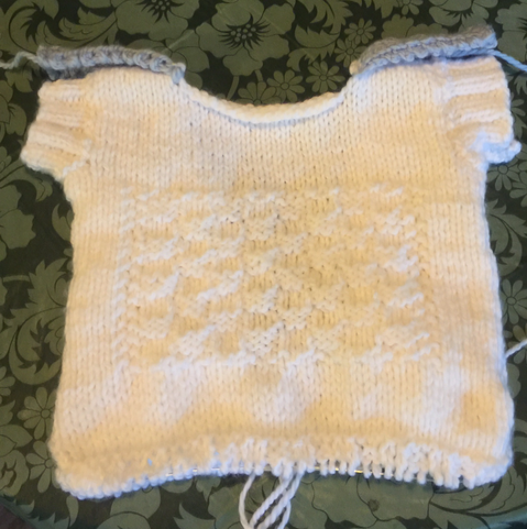 The same white sweater vest, on a green tablecloth background.  Pictured is the back, showing the checkerboard pattern knit into the design. The bottom stitches are still live, seen on a plastic knitting needle wire, where the ribbing at the waist will be added.