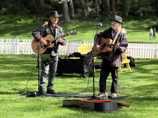 Two older men standing on a grass field holding guitars in front of microphones.