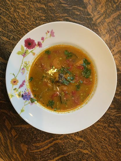 We have a round white dish. We have a thin red broth with diced vegetables. Green parsley accents the meal. The background is a table of dark wood