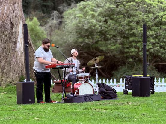 One keyboardist and a drummer on a grass field in front of some trees and bushes.
