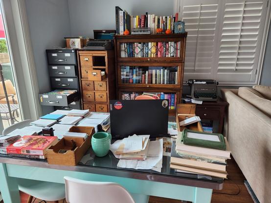 A busy desk with a black laptop on one side, but surrounding it are a variety of piles of 2-3 and up to 100 index cards each. We also see two wooden card index boxes including one which has been taken from one of the two card index filing cabinets standing behind the desk.