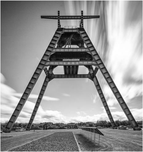 A large, metal mining tower structure is featured in the image, set against a cloudy sky. The tower has an A-frame design and is surrounded by a gravel path and displays. The image is in black and white.