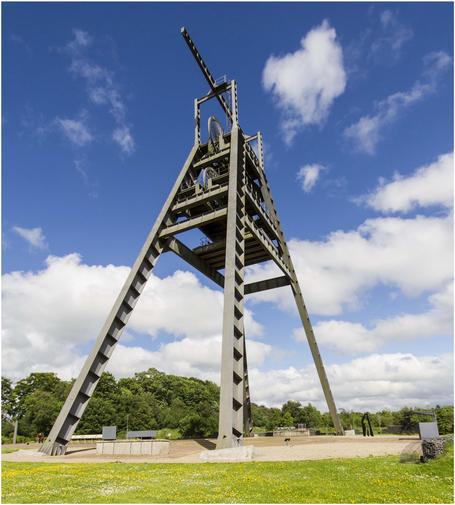A tall industrial structure resembling a mining headframe with a metal framework, set against a blue sky with fluffy clouds. Surrounding greenery and a flower-filled ground are visible.