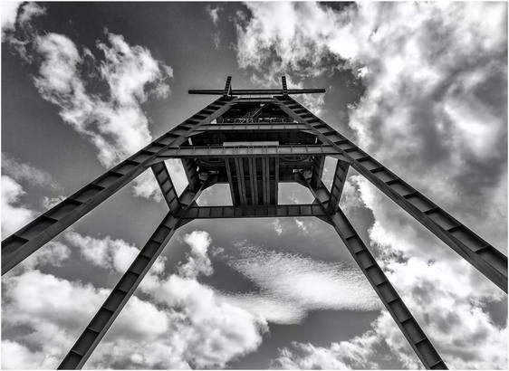 A black and white image of a tall industrial structure or tower, viewed from below, against a cloudy sky. The stark lines of the metal framework contrast with the softness of the clouds.