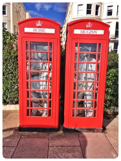 Two old style BT red telephone boxes side by side with what appears to be two mannequins dressed in grey hazmat suits inside. The phone boxes have the name Rosie and McGinn above them and are set on the pavement against a hedge and some tall apartments behind them.