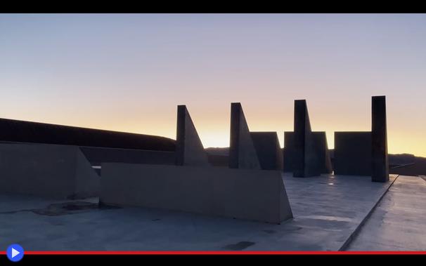 Strana composizione scultorea con triangoli e pannelli di cemento si staglia contro il sole dell'alba nel deserto. Il sito sembra estendersi ben oltre l'area contenuta nell'inquadratura.