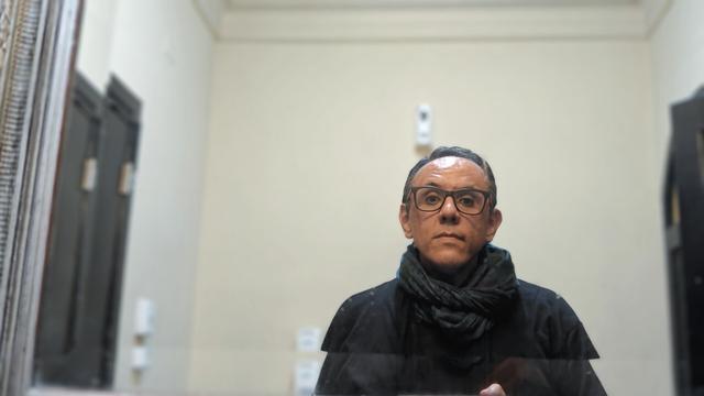 Un hombre se mira en un espejo apaisado. Es moreno, tiene cabello negro y viste del mismo color. Apenas se ve el teléfono con cuya cámara se toma la foto. Al fondo la pared es blanca.
