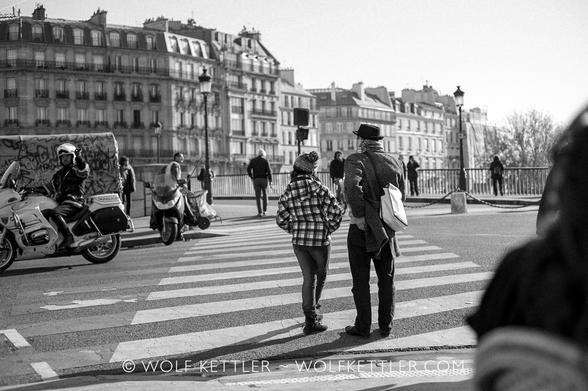 In this black & white photograph, a zebra crossing leads away from the camera towards a bridge,  Pont Saint-Louis in Paris, France. In the foreground, seen from behind, a father and his teenage daughter are waiting to cross the road.
Across the road in the middle distance on the left margin of the frame is a policeman on a motorbike. He is holding his arm up.