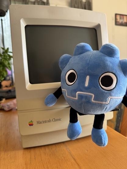 a plushie shaped like the Godot fame engine logo (a blue gear-shaped robot head) sitting on top of a floppy disk that‘s partially inserted in a Macintosh Classic