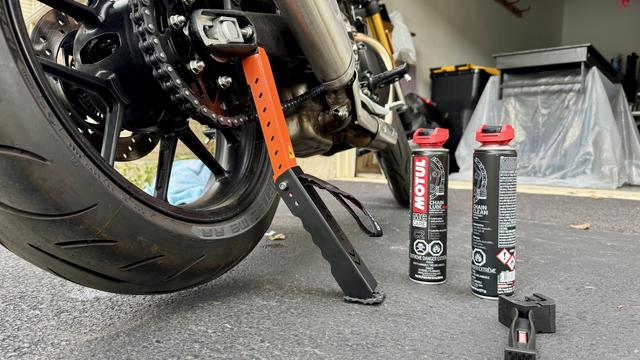 A motorcycle's rear wheel and chain are visible, with a black and orange paddock stand supporting the bike. Two cans of Motul chain lube and chain cleaner are positioned nearby, along with a smaller tool on the ground.