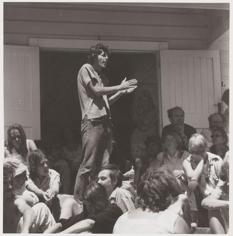 The image is a black and white photograph capturing a moment during an outdoor gathering, likely related to anti-Vietnam war protests. A central figure stands on what appears to be a small elevated platform or step in front of several seated individuals who are attentively listening.

This person gestures with their hands open as if explaining something passionately or addressing the crowd directly. The individual is dressed casually in a short-sleeved shirt and jeans, suggesting an informal atmosphere typical for such demonstrations.

The audience consists mostly of younger adults with varied expressions—some appear engaged while others look more contemplative or disinterested. There's no clear indication of exact numbers due to the angle from which the photo was taken; however, it seems like a group not too large but substantial enough to suggest collective action and shared purpose.

Behind them stands another person who appears to be taking notes on paper held in hand, hinting at documentation or reporting activities. The setting is casual with visible wooden structures that could indicate this event took place near community centers or houses of residence rather than official public spaces.

The composition focuses mainly on the speaker's gesture and audience engagement but also gives a glimpse into their environment—outdoors during daylight hours in what appears to  [...]