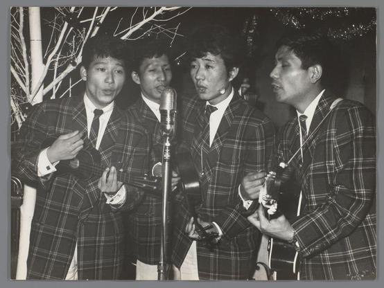 The image is a black and white photograph capturing four individuals, seemingly engaged in musical performance. They are dressed in matching dark plaid suits with ties, which suggests they might be part of a group or band. Each person holds a stringed instrument: two have ukuleles, while one has what appears to be an acoustic guitar held in their hands. The other individual is holding onto a microphone stand but not actively singing into the mic.

The background features bare branches and some foliage that indicates it could be taken during winter or late fall when trees are devoid of leaves. It's night time as suggested by the darkness surrounding them, with artificial lighting possibly highlighting the group for their performance.

This photo seems to capture a candid moment from what might have been an outdoor public event in Tokyo's Asakusa district, given the context provided on Vier Japanse zangers (Four Japanese Singers) and Westerse kleding (Western clothing). The attire of the singers is Western-inspired but with Asian influences. This blend suggests cultural fusion or perhaps a specific style chosen for their performance.

The historical attribution to Ed van der Elsken indicates that this photograph was taken during his tenure capturing various aspects of Tokyo's life and culture, likely between 1959-60 based on the provided reference link.