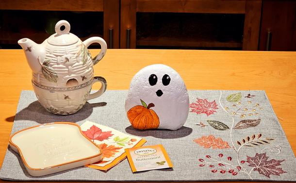 A simple Autumn tea tableau on a grey linen mat embroidered down the right side with leaves in somber Fall colors. In the upper left is a ivory ceramic tea-for-one set shaped like a woven basket, painted in soft tones of sage & fawn with pinecones, acorns, dragonflies & bees. In front of it is a ceramic plate in the shape of a slice of white bread with a brown crust. There are a Fall-themed napkin & a packet of Twinings Buttermint tea beside the toast. In the center of the mat, standing up, holding an orange pumpkin, is a white ghost with a look of surprise on its sweet face. The ghost is actually a large rock fully painted to be the three- dimensional ghost.