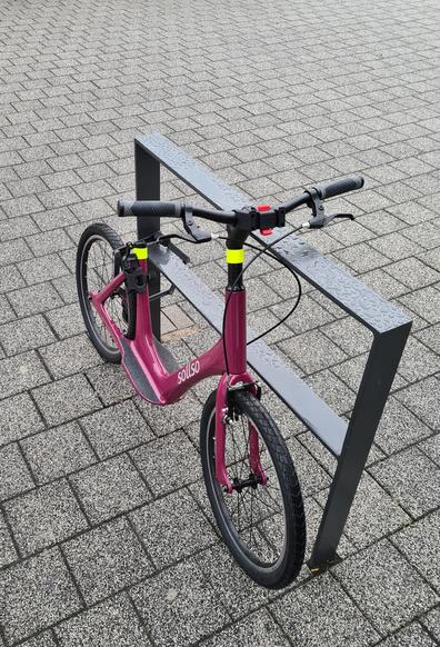 Auf einem grau gepflasterten Boden ist im unteren Bildbereich, mittig, leicht links ein metallenes, dunkelgraues, rechteckige, Zaunelement. 
An diesem ist ein Laufrad für Erwachsene befestigt. Durch das Hinterrad, den Rahmen und das eben beschrieben Element wurde ein Klapp-Schloss geführt. Die Räder haben zwanzig Zoll.
Es gibt rutschfeste Abstellflächen für die Füße, recht und links am Rahmen.
Das Rad ist dunkel Pink, beerig gefärbt. Es wurden zusätzlich neon gelbe Reflektoren mittig am Lenker und an der Sattel-Stange befestigt.
Der Sattel fehlt.