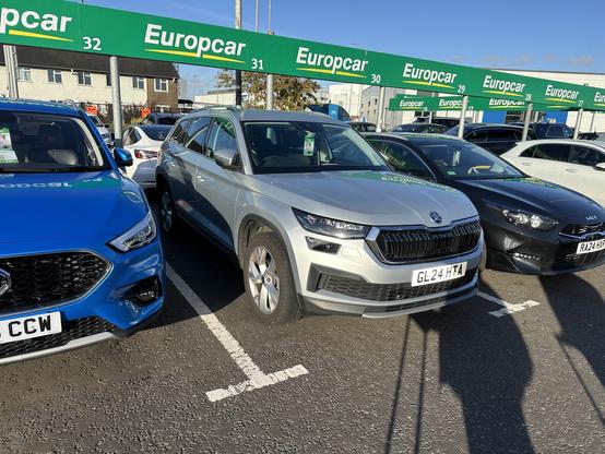 A parking lot with several cars, including a blue vehicle on the left, a silver SUV in the center labeled with the registration "GL 24 HTA," and a black car on the right. The Europcar signage is visible in the background.