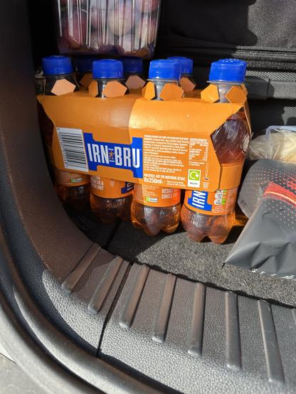 A pack of eight Irn-Bru soft drink bottles is shown, each 250ml, placed in the trunk of a car. The background includes a container with fruits and other items.