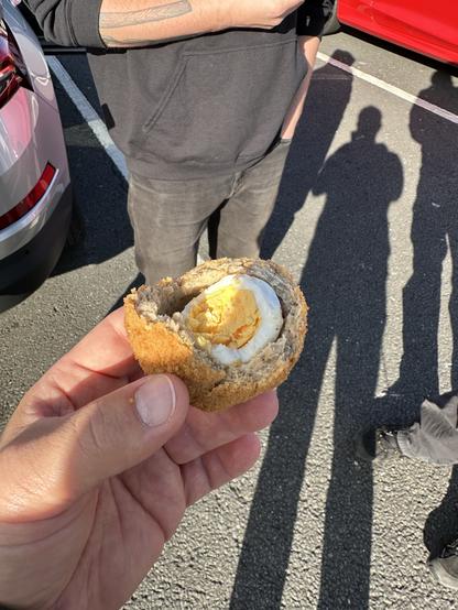 A hand holds a half of a scotch egg, showing the soft-boiled egg inside, with two people in the background. The setting appears to be a parking lot.