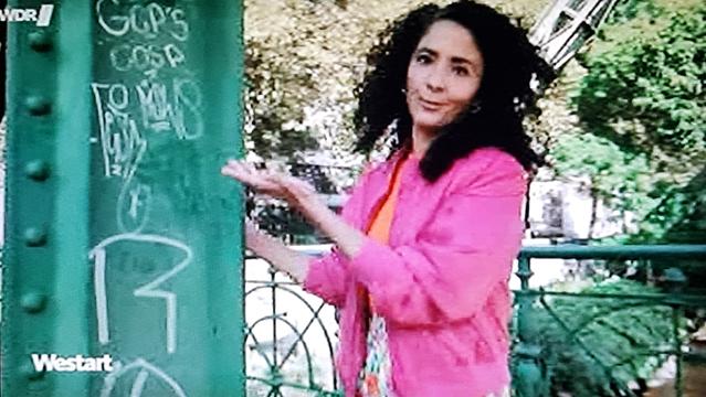 Fernsehansagerin in Rosa zeigt auf grünen Brückenpfeiler
