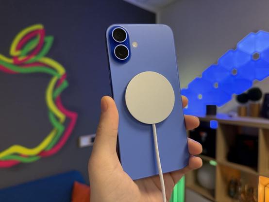 Eine Hand hält ein blaues iPhone 16 samt angelegtem MagSafe-Qi2-Ladekabel von Apple in die Kamera. Im Hintergrund ist unscharf ein Raum mit Apple-Logo an der Wand, einem Regal und blauer Nanoleaf-Beleuchtung zu sehen.