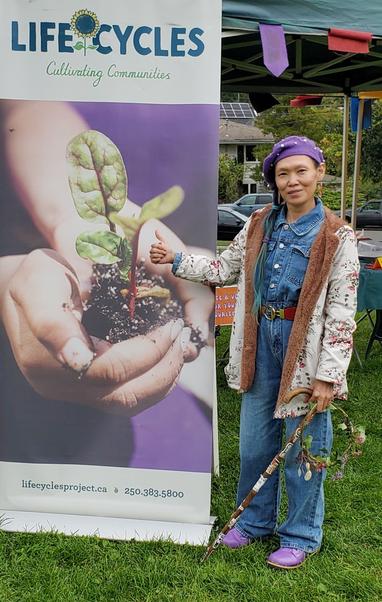 Me standing beside LifeCycles Project banner sign - it has photo of hands holding Swiss chard seedling.