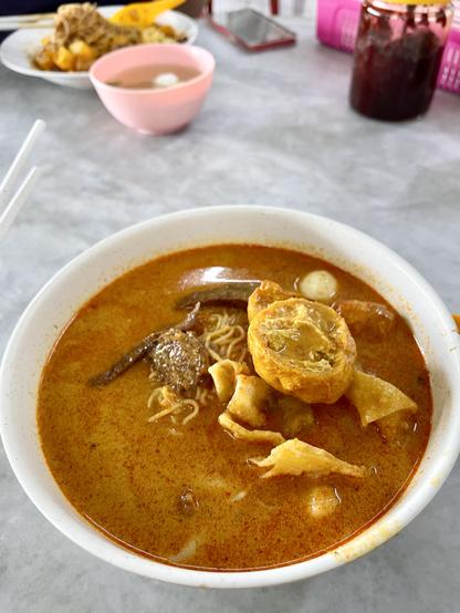 A bowl of wheat noodles in curry gravy. There are fried wanton dumplings, tofu puffs and some other ingredients as well.