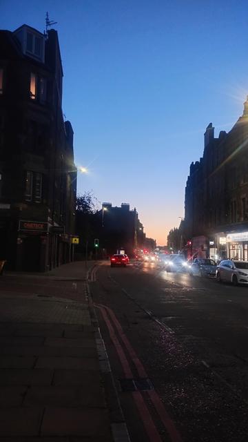 Post sunset sky visible above the road between the tall, Victorian stone tenements, the colours shading from darker blues to lighter near the bottom, and a yellow and orange band at the horizon
