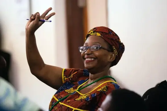 Stella Nyanzi, a black woman smiles at the camera, raising her right arm with two fingers in a victory sign