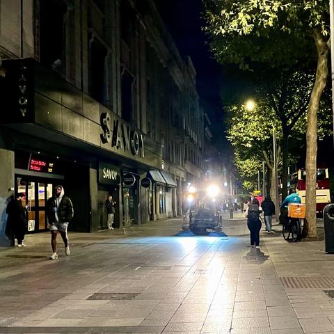 A nighttime scene of a city street featuring the Savoy cinema. People are walking along the wide pavement, and there is a vehicle with headlights on in the background. Trees line the street, with urban elements like rubbish bins and bicycles visible.