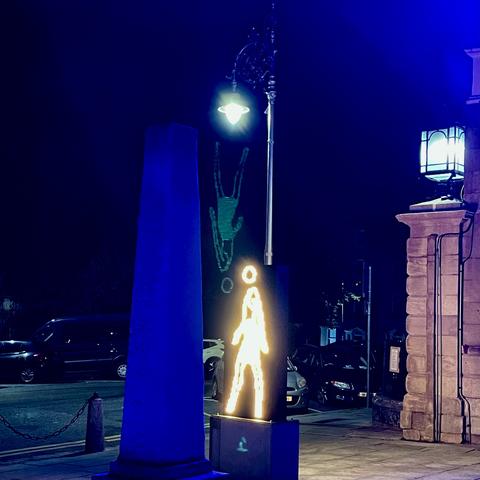 A glowing figure sculpture illuminated in white, set against a dark background with blue lighting. The scene includes a pillar and a streetlamp, as well as cars parked nearby. A faint green reflection of a person diving is visible on the wall behind the light