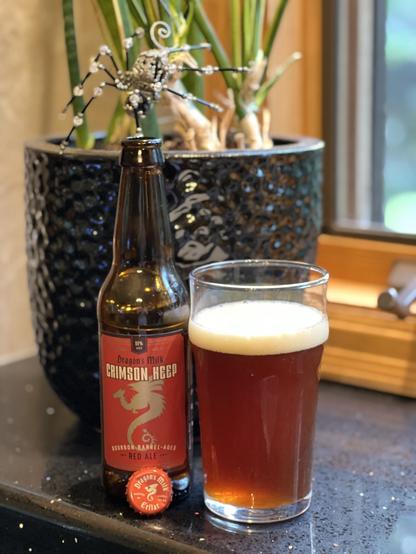 A pint glass of deep red ale stands next to a brown bottle with a dark red label with white lettering and a gold dragon in silhouette. A spider hangs above the bottle on a plant