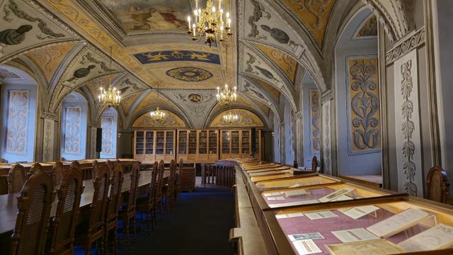 Hall named after Pranciškus Smuglevičius in Vilnius University: Cassicism style interior with arcs, long wooden table with chairs, large book shelves in the back, backlit exposition of books under glass, large chandeliers