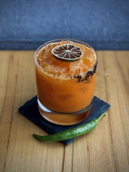 bright orange cocktail with dehydrated lime garnish and black salt rimmed old fashioned glass sitting on a black slate coaster and beechwood table