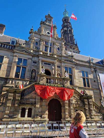 Close up of the frontage of Leiden city hall, with the decoration and flags in red and white, the colours of the city flag. Two statuettes are holding small flags of their own. A person with long blond hair and a festive jacket is cycling past.