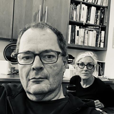 Candid black & white photo of my husband Alan and me, seated in our kitchen with cabinets and cookbook shelf behind us.