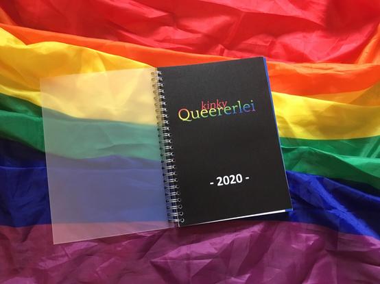 Ein Ringbuch mit offenem Plastik-Schutzdeckel. Der Titel ist „kinky Queererlei 2020“. Das Buch liegt auf einer teilweise zerknüllten Regenbogenflagge.