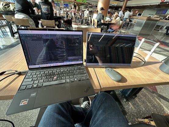 A workspace in an airport features two screens on a wooden table. The main laptop displays coding software, while a secondary monitor shows similar content. A mouse is positioned nearby, and a suitcase can be seen beneath the table. People are seated in the background, eating. 

I’m not, and now I’m hungry.
