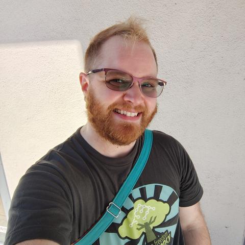 White person with a blonde faux-hawk, a red-brown beard, and purple glasses. They are wearing a gray t-shirt with the Software Freedom Conservancy logo on it.