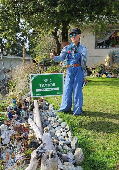 Me, in my wide leg denim jumpsuit, on our front lawn, giving a thumbs up, beside a BC Greens Saanich South, Ned Taylor, support sign.