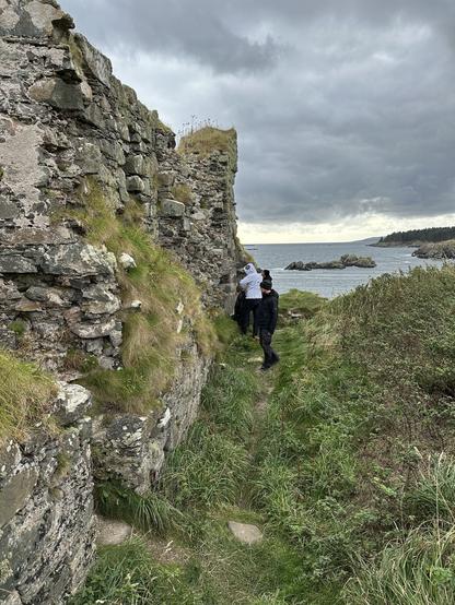 Two individuals stand near the remnants of a stone wall by the coast. The scene features a grassy pathway leading to the water, with a cloudy sky overhead.