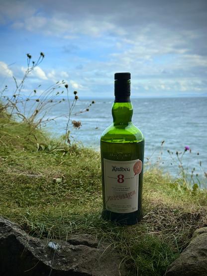 A bottle of Ardbeg 8 Year Old Islay Single Malt Scotch Whisky is placed on grass and rocks, with a picturesque view of the ocean and cloudy sky in the background.