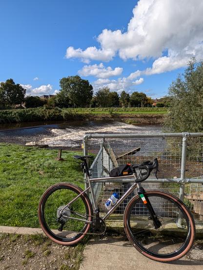 The image shows a gravel bike parked near a river. The bike is a North Road Cycles Grav-i-Ti, which is a titanium frame gravel bike. The bike has a silver frame, brown handlebar tape, and brown tyres. The river is flowing fast and there is a weir in the background. The sky is blue with white clouds.